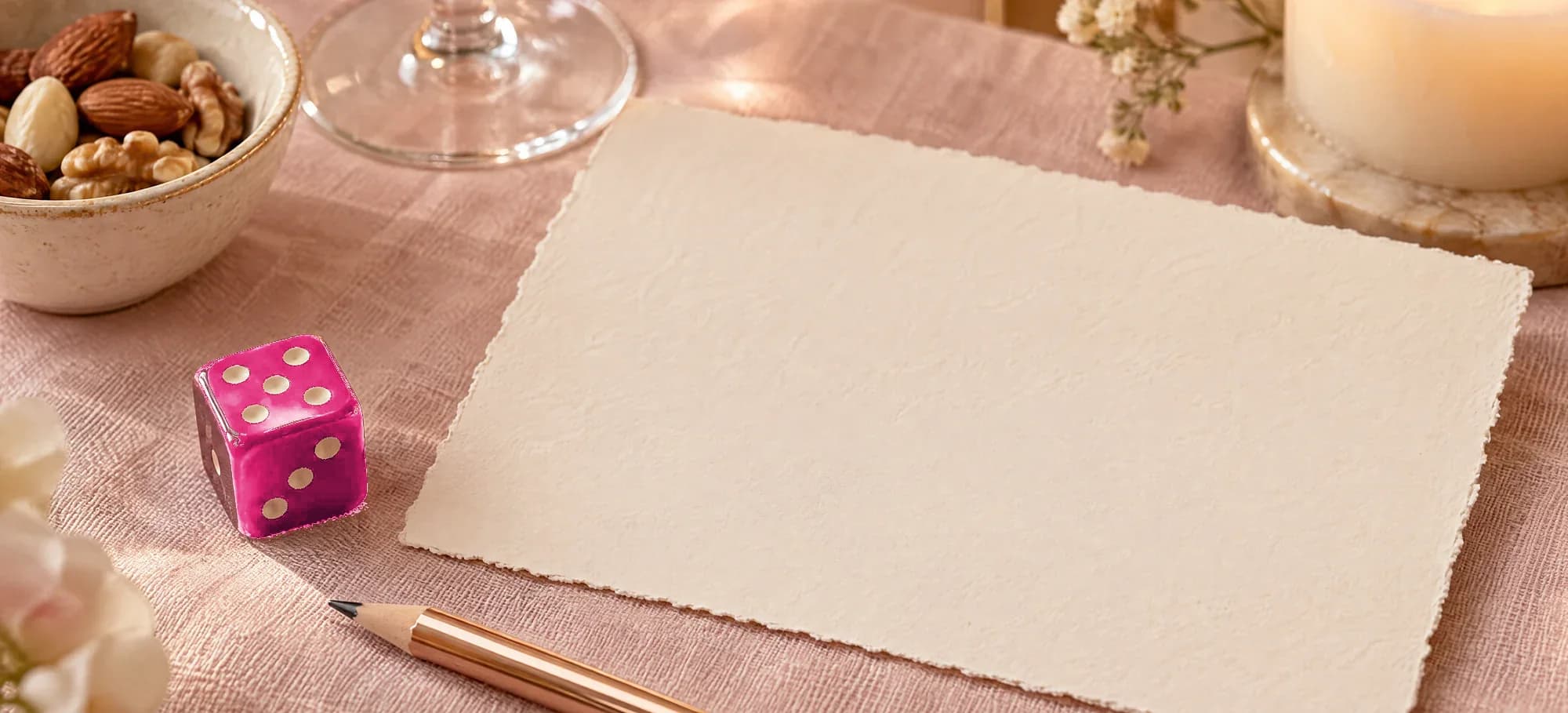 A blank deckle-edged card on blush linen, with a pink die, a rose-gold pencil and a lit candle beside it.
